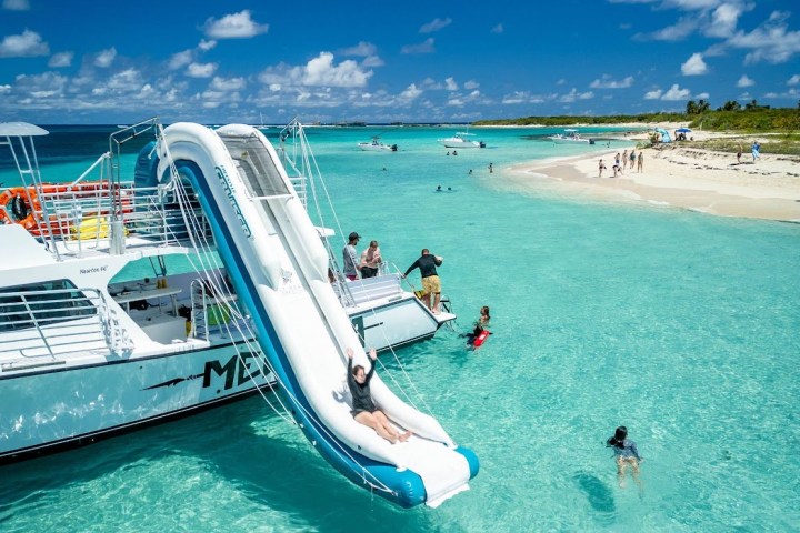 Catamaran with slide into clear turquoise water by sandy beach.