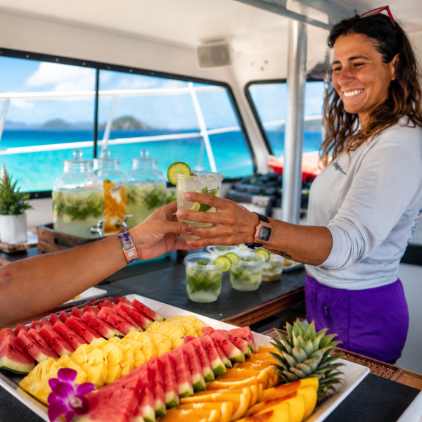 Smiling woman serves drinks on boat with tray of colorful fruit.