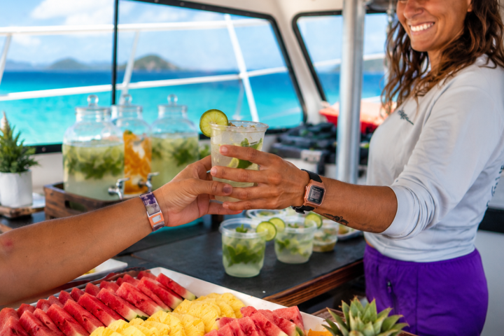 Smiling woman serves drinks on boat with tray of colorful fruit.