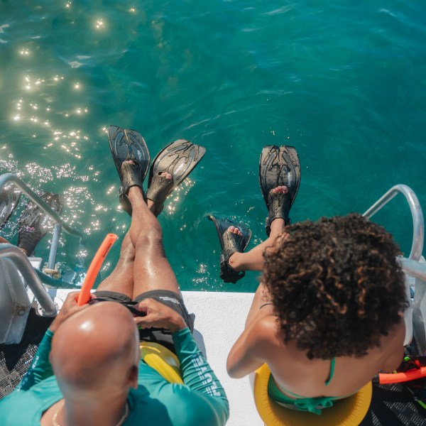 Two people in swimwear and fins sit on a boat edge over clear water.