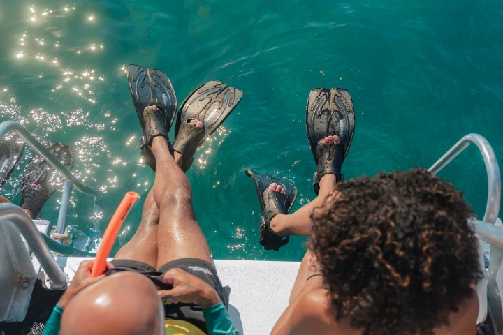 Two people in swimwear and fins sit on a boat edge over clear water.