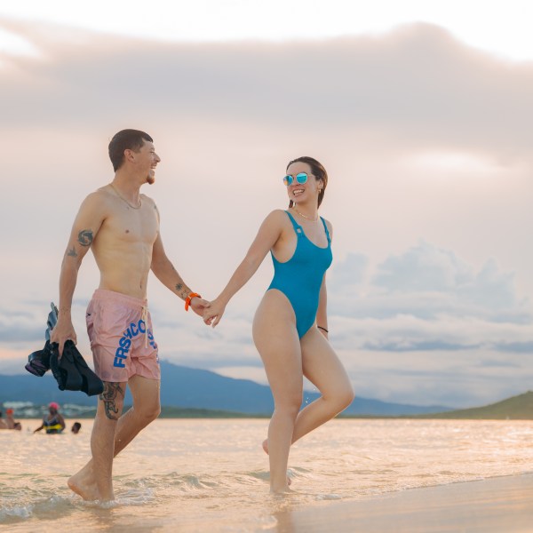 Couple holding hands, walking in shallow water at the beach, with mountains in the background.