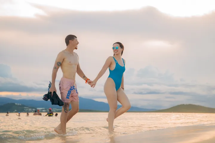 Couple holding hands, walking in shallow water at the beach, with mountains in the background.