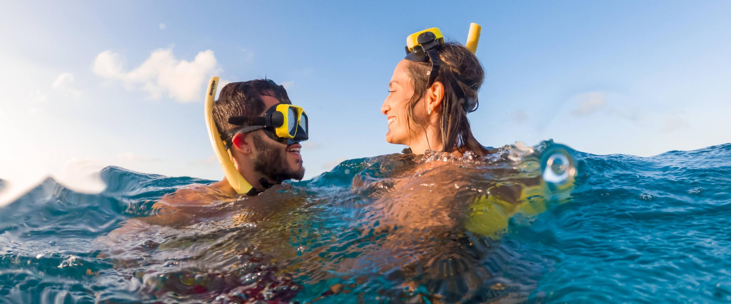 Two people snorkeling in blue water, smiling and facing each other under a clear sky.