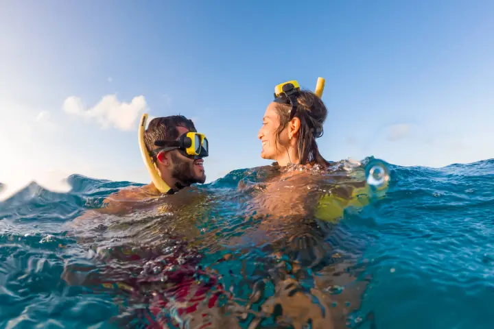 Two people snorkeling in blue water, smiling and facing each other under a clear sky.