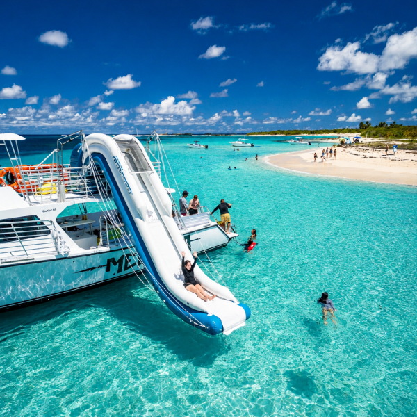 Catamaran with slide in clear blue water, people swimming near sandy beach.
