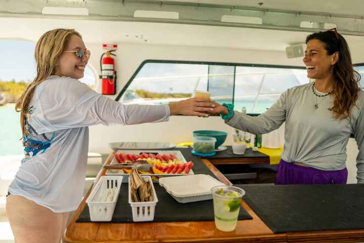 Two people smiling and exchanging drinks on a boat with a food spread.