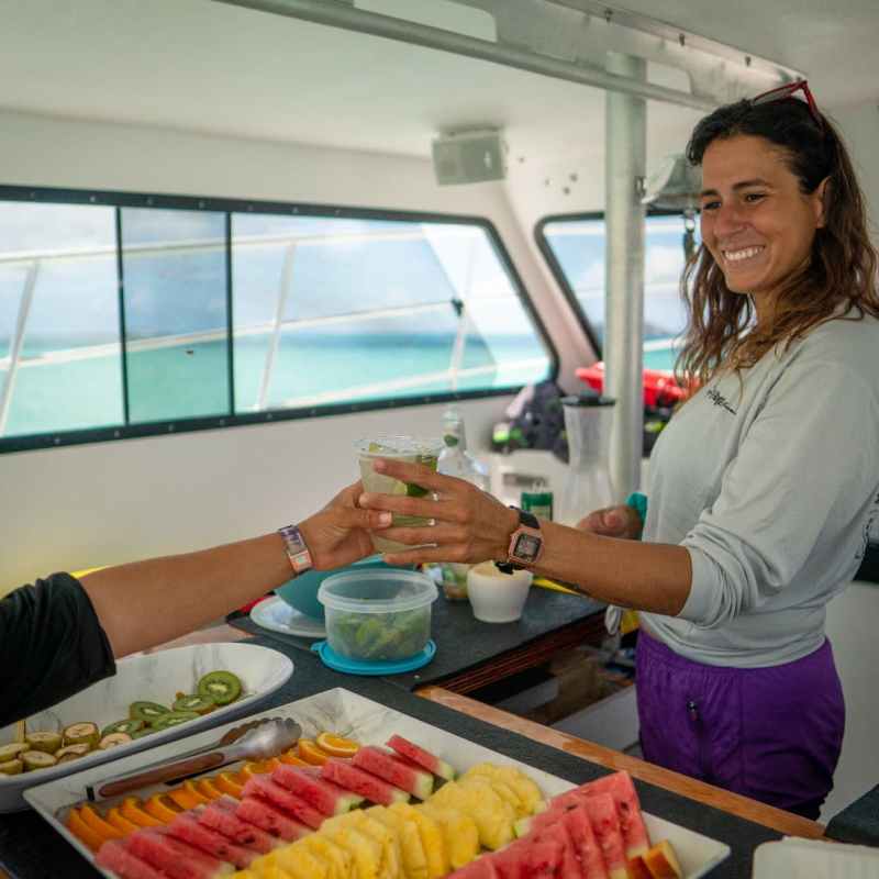 Smiling person on a boat serving a drink beside a tray of sliced fruit.