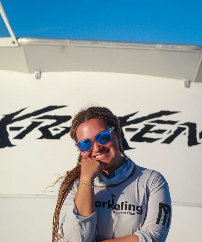 Person smiling in sunglasses and braid, wearing a snorkeling shirt with the blue sky background.