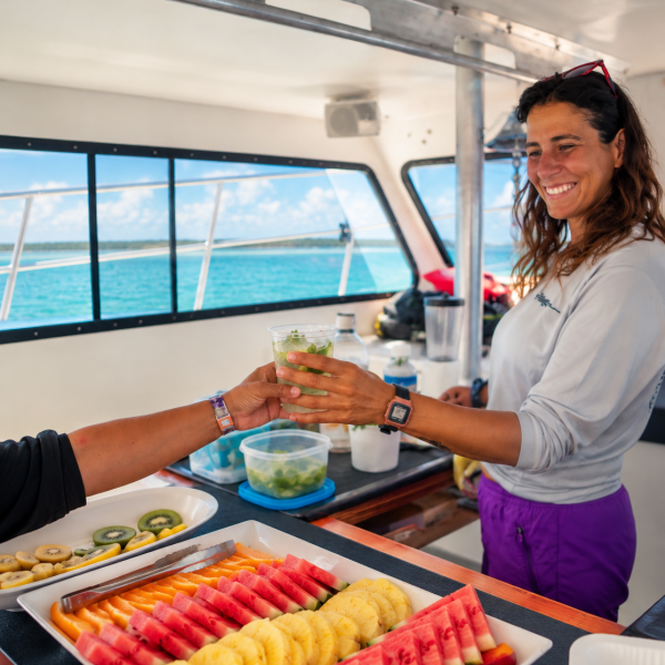 Woman on boat serving drink beside a tray of assorted fruit.