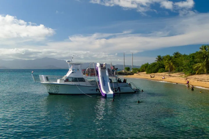 Boat with waterslide near tropical beach, clear blue water, and palm trees.