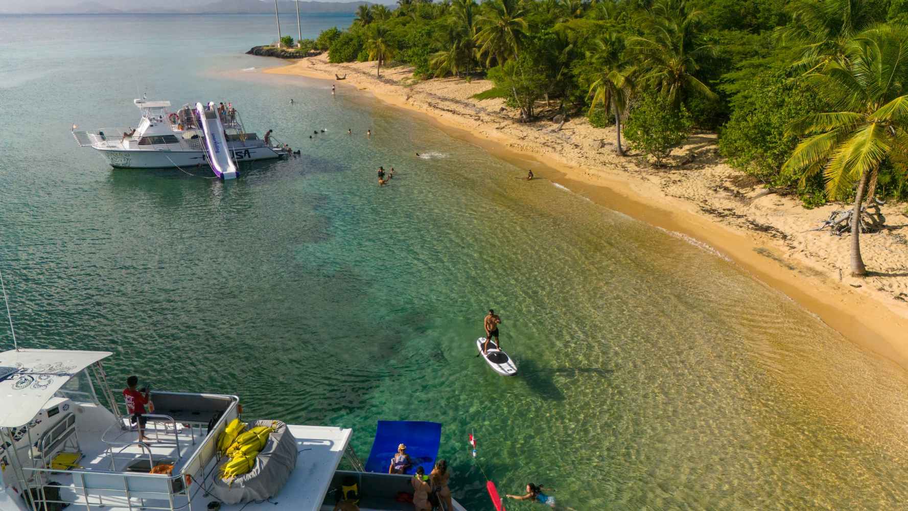 Boats near a tropical beach with people swimming and paddleboarding.