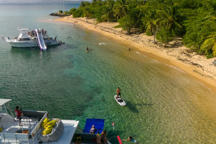 Tropical beach scene with boats, people swimming, and palm trees along the shoreline.