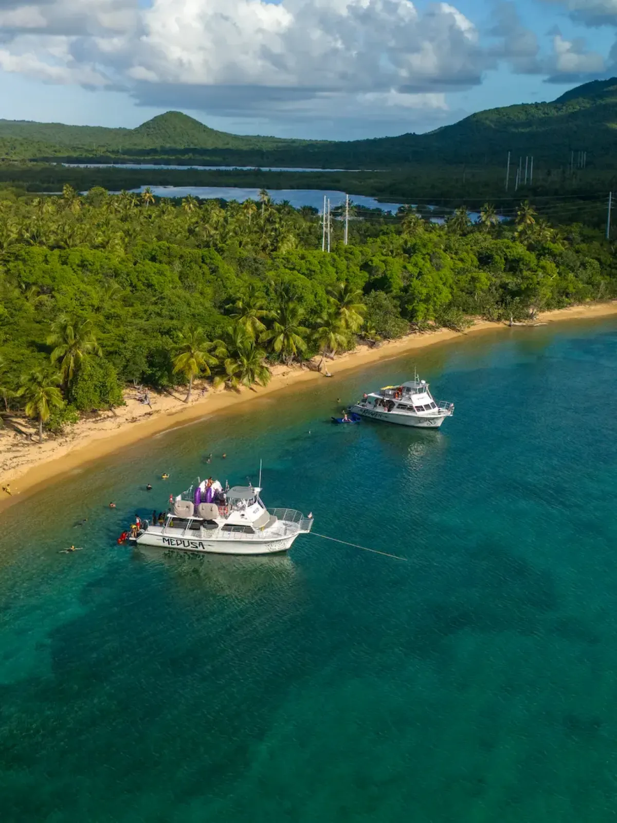 Two boats anchored near a lush, tropical beach with a mountainous backdrop.