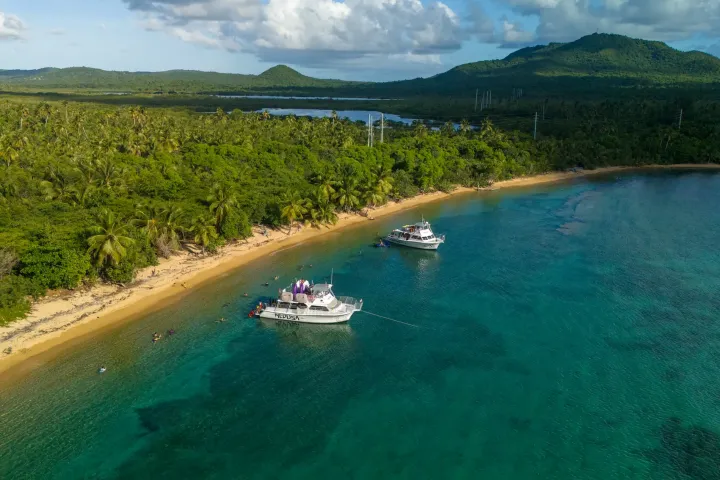 Two boats anchored near a lush, tropical beach with a mountainous backdrop.