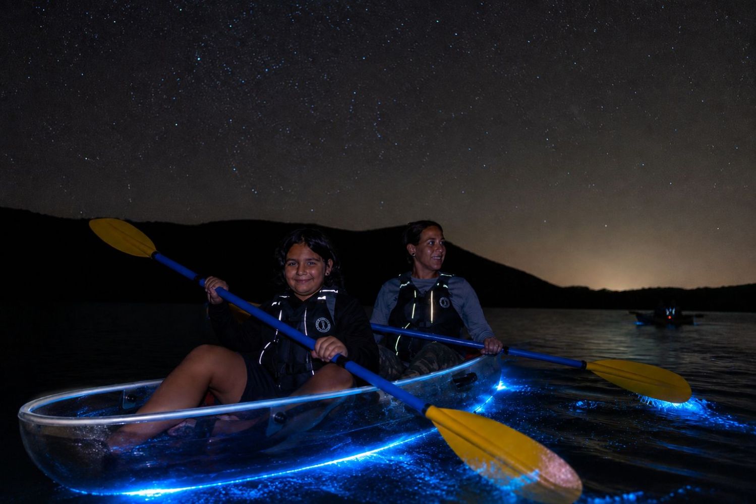 People paddle transparent kayaks glowing at night on a starry lake.