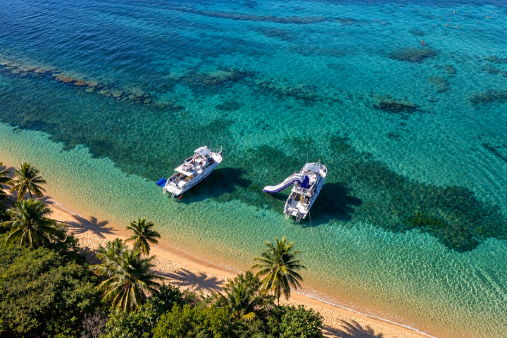 Two boats with slides on turquoise water near a sandy beach with palm trees.